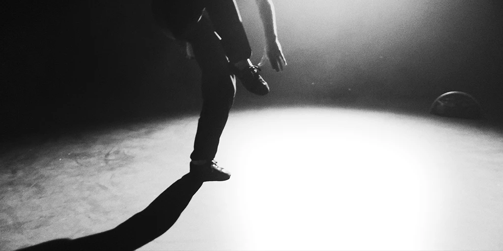 Es ist ein ovaler weißer Lichtkegel auf dem Boden zu sehen. Im Lichtkegel tanzt ein junger Mann. Es sind seine Beine und sein linker Arm zu sehen. Sein rechtes Bein ist stark angewinkelt. Er trägt eine schwarze Hose. Rechts im Hintergrund steht ein Bluetooth Lautsprecher.