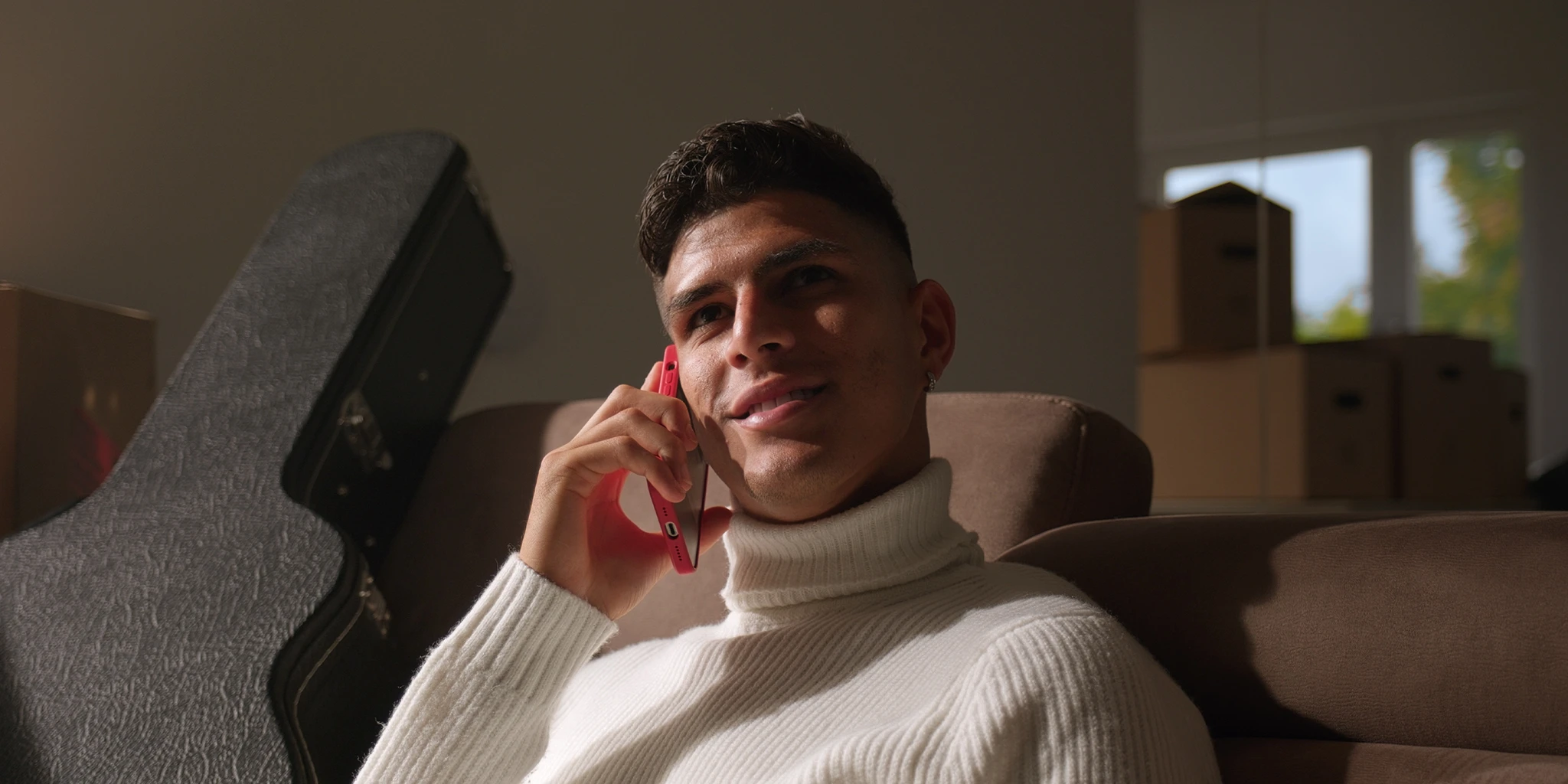 Ein junger Fußballer, Piero Hincapié, sitzt auf seinem Sofa und telefoniert. Er trägt einen weißen Pullover. Neben ihm steht ein Gitarrenkoffer und im Hintergrund sind Umzugskartons zu sehen.