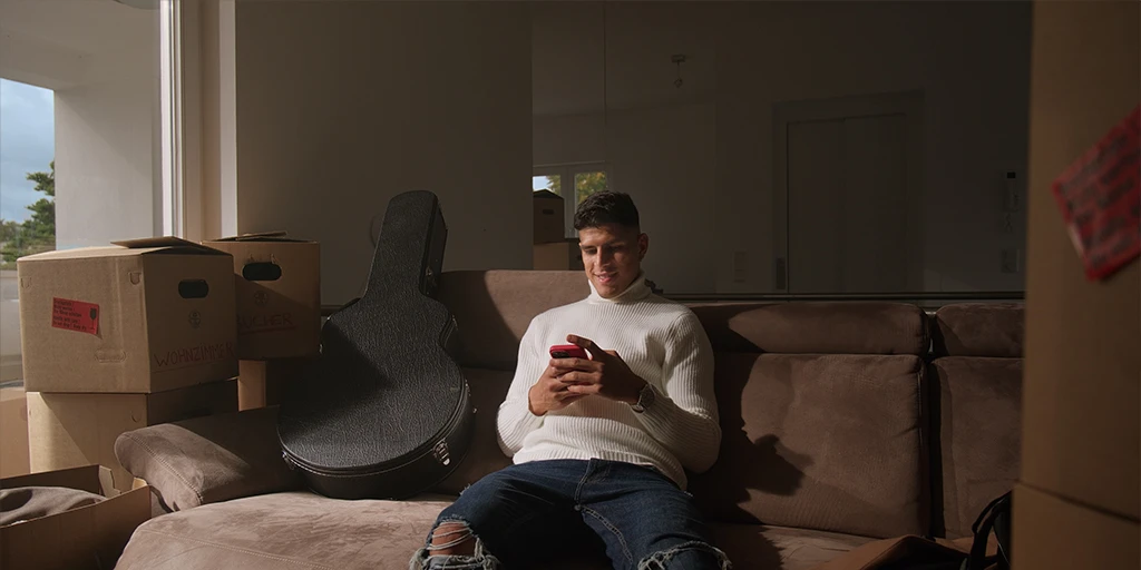Ein junger Fußballer (Piero Hincapié) sitzt auf seinem Sofa und tippt auf seinem Handy. Er trägt einen weißen Pullover. Die Sonne scheint durch die Balkontüre. Neben ihm stehen Umszugskartons.