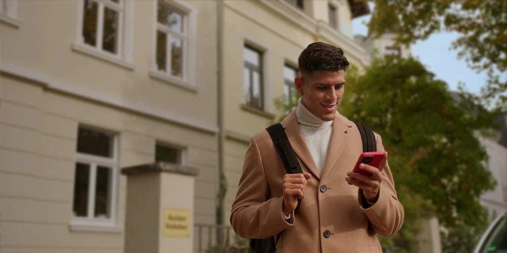 Ein junger Mann (Piero Hincapié) trägt einen Mantel und schaut auf sein Handy. Er trägt einen Rucksack und steht vor einem gelblichen Haus.