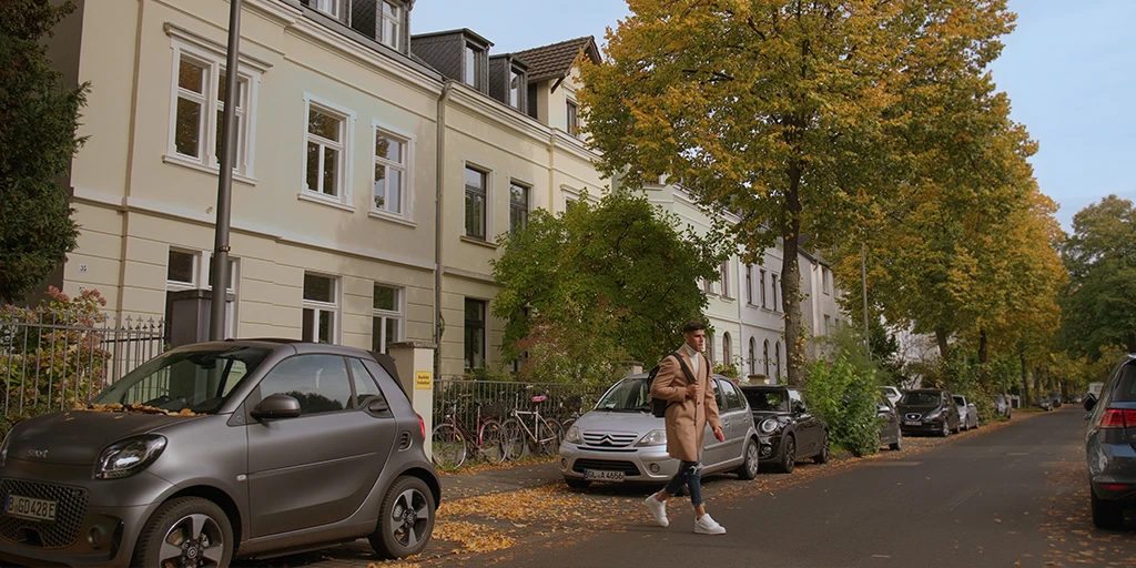Ein junger Mann (Piero Hincapié überquert eine Straße. Er trägt einen braunen Mantel. Hinter ihm befindet sich ein gelbliches Haus. In der Flucht ist eine Allee mit herbstlichen Bäumen zu sehen.