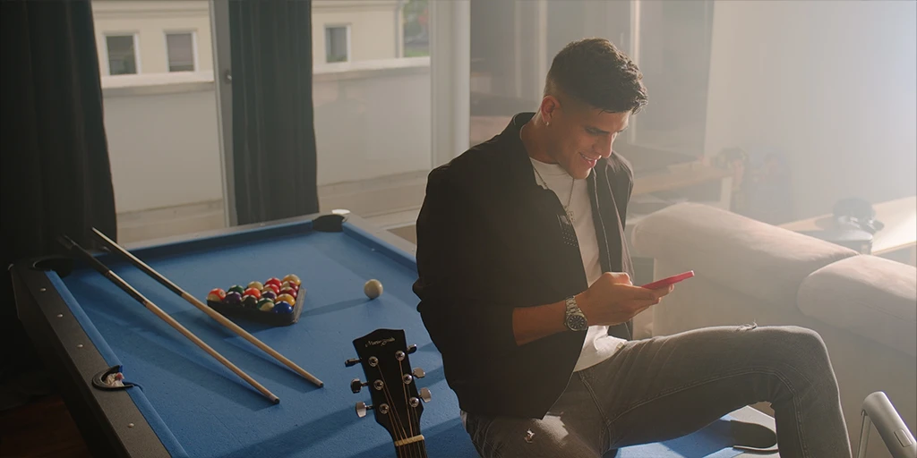 Ein junger Mann (Piero Hincapié) sitzt in seiner Villa auf einer blauen Billiard Platte. Er hält ein Handy in seiner Hand. Der Raum wird von Sonnenlicht durchflutet. Neben ihm sieht man eine Gitarre.