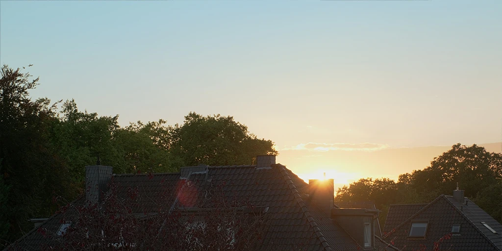 Die Sonne geht unter und verschwindet hinter einem Schornstein von einem Wohnhaus. Es sind zwei Dächer zu sehen. Dahinter befindet sich ein Wald. Der Himmel weist einen blau-rötlich-gelben Farbverlauf auf.