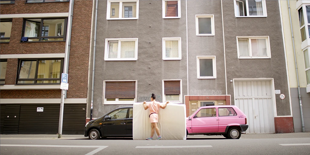 Die Protagonistin des Films »Die Mathematik der Dinge« transportiert eine Matratze. Sie trägt ein pinkes Kleid und läuft vor einem pinken Auto entlang. Im Hintergrund ist das Haus von dem Mann zu sehen, der ihr die Matratze geschenkt hat.
