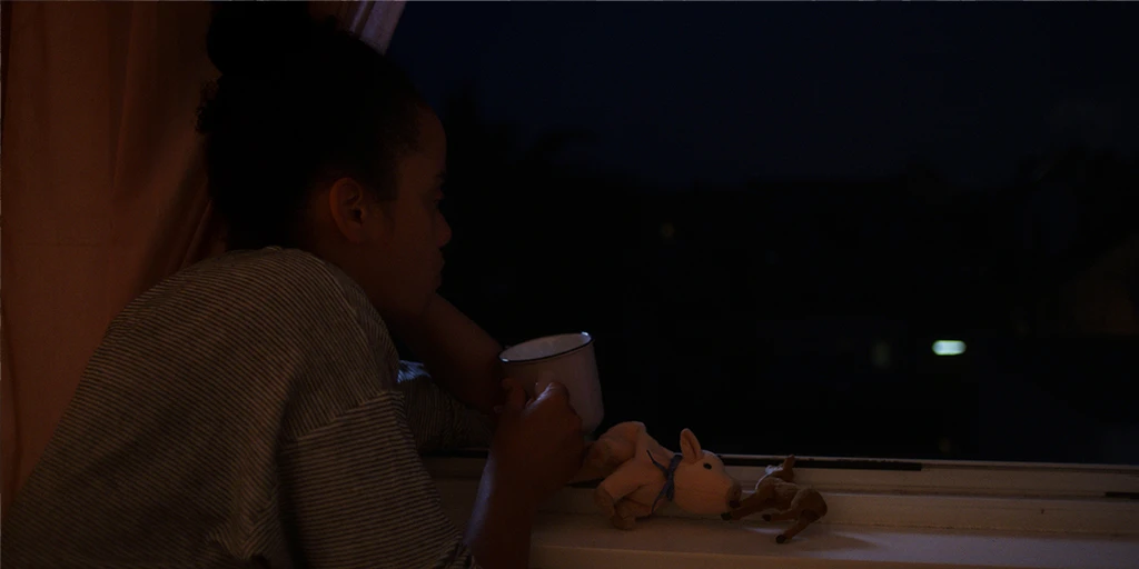 Eine junge Frau (Shari Asha Crosson) sitzt nachts am offenen Fenster mit einer Tasse Tee. Vor ihr liegen zwei Kuscheltiere. Sie schaut nach Draußen.