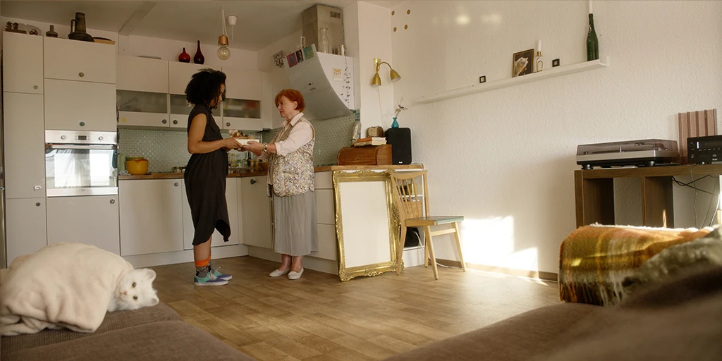 Eine ältere Dame (Margit Laue) überreicht in ihrer Wohnung der Protagonistin Lena (Shari Asha Crosson) des Films »Die Mathematik der Dinge« eine Schachtel mit Kuscheltieren. Eine Katze liegt auf dem Sofa und starrt in die Kamera. Der Raum ist sonnendurchflutet.