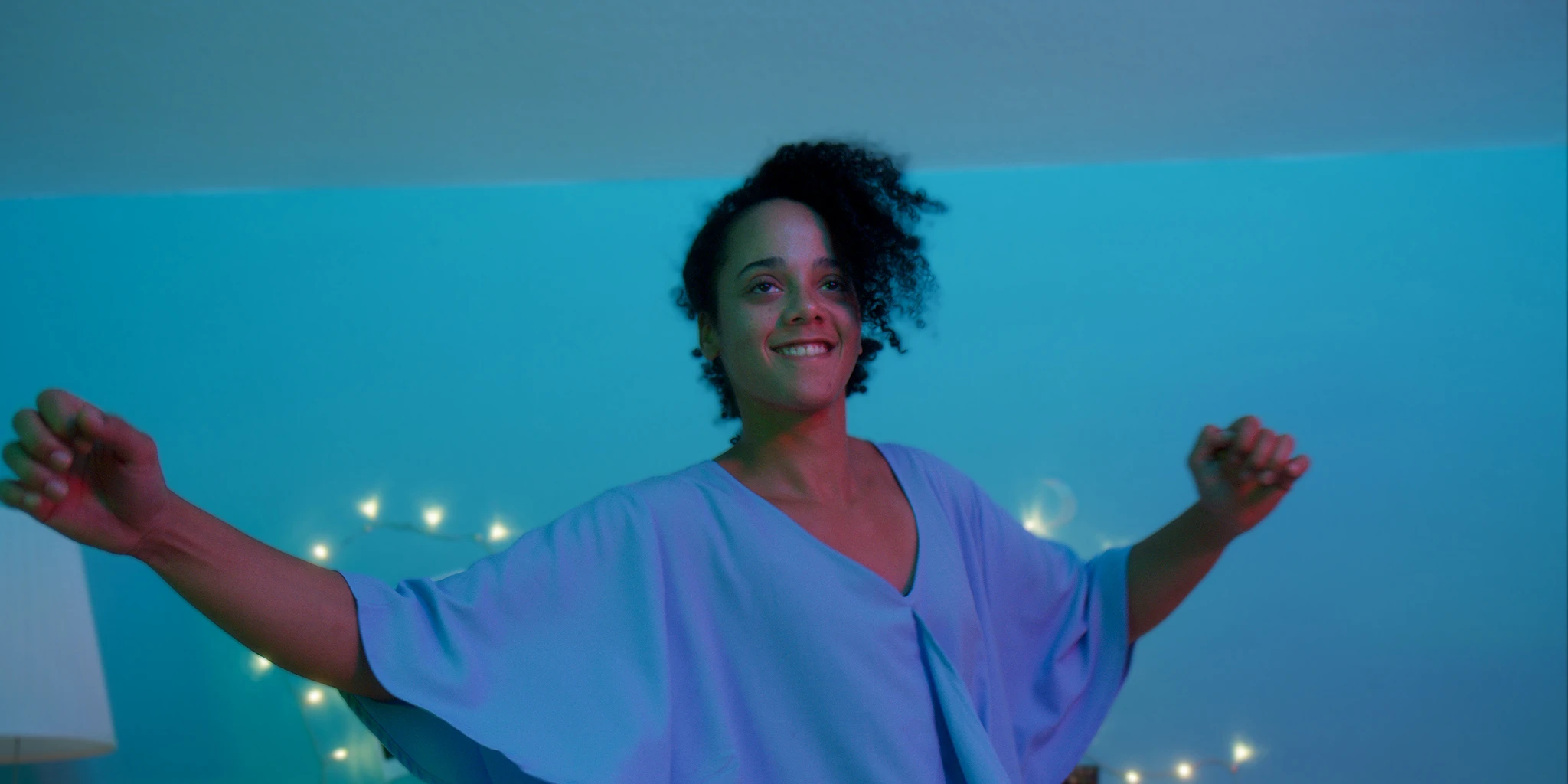 Eine junge Frau (Shari Asha Crosson) tanzt offen und ausgelassen vor eine blau-grünen Wand. Im Hintergrund ist eine Lichterkette.