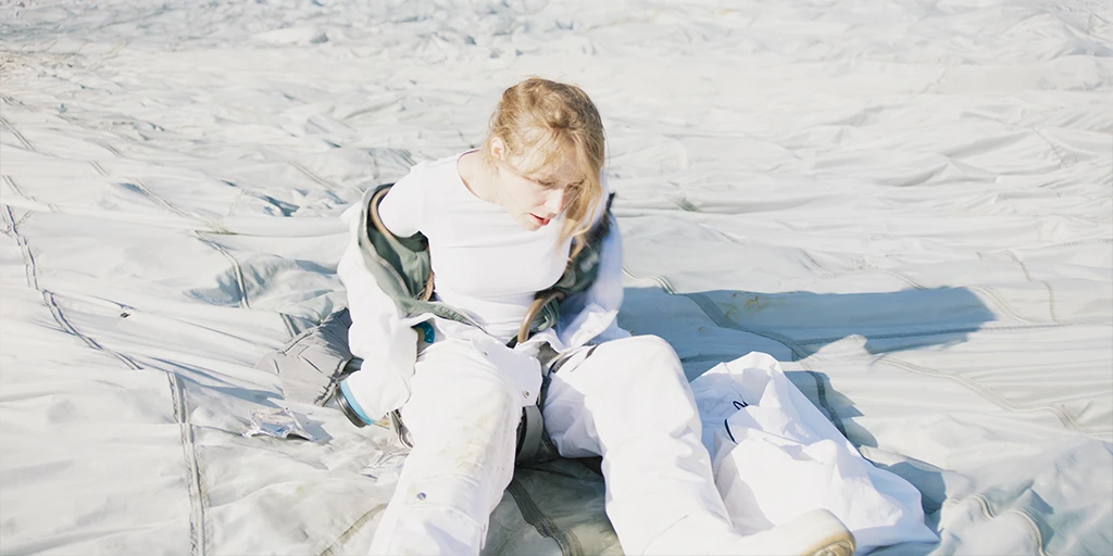 Eine Astronautin (Odine Johne) sitzt auf einem weißem Lastenschirm. Mühsam pellt sie sich aus Ihrem Raumanzug im Spielfilm »Im Meer der Stille«.