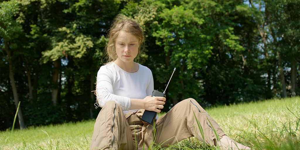 Eine Astronautin (Odine Johne) hält ein Funkgerät in ihrer Hand und blickt ins Gras. Sie trägt ein weißes Top und eine braune Hose im Spielfilm »Im Meer der Stille«