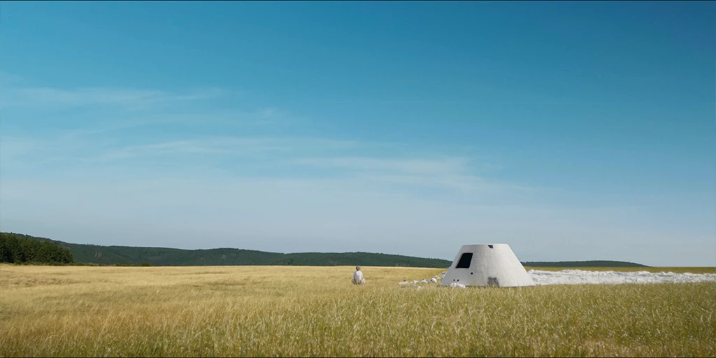 Die Astronautin (Odine Johne) sitzt neben ihrer Raumkapsel und ihrem Lastenschirm im Gras und blickt in die Ferne einer Graslandschaft. Am weit entferntem Horizont bildet sich ein Wald ab im Spielfilm »Im Meer der Stille« © Johannes Schmülling