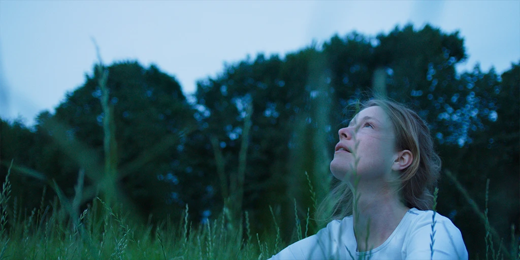 Eine Astronautin (Odine Johne) sitzt im Gras und schaut in das Universum. Vor einem bläulichem Abendhimmel weht leichter Wind durch ihr Haar. Sie trägt ein weißes Top im Spielfilm »Im Meer der Stille«