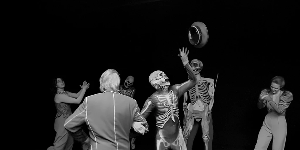 Ein dunkler Raum. Direkt vor uns tanzt ein Mann gehobenen Alters und ein Skelett Arm in Arm im Kreis. Das Skelett macht sich einen Spaß daraus, einen Hut in die Höhe zu werfen. Im Hintergrund klatscht eine Frau in die Hände. Eine weitere Person ist in einem Skelett-Kostüm. Rechts davon ist eine Skelett-Puppe zu sehen, die von einer Puppenspielerin gesteuert wird. Rechts davon ist eine junge Frau zu sehen, die amüsiert in klatscht.