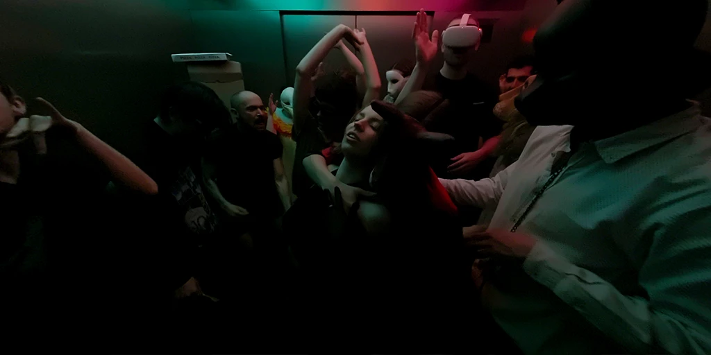 Wir stehen in einem großen, sehr gut gefüllten Aufzug. Die überwiegend junge Partygesellschaft tanzt ausgelassen. Eine junge Raverin tanzt mit einem Mann mit Hundemaske. Ein großer Mann trägt einen VR Brille. Der Aufzug ist atmosphärisch, nur durch ein grün-magentafarbendes Deckenlicht beleuchtet.
