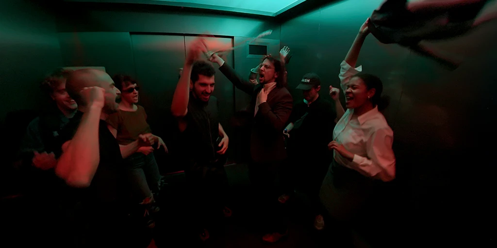 Acht Menschen tanzen in einem Halbkreis in einem großen Aufzug. Grün-oranges Licht trifft sie von oben. Eine junge Frau im business Outfit schleudert eine Jacke durch den Aufzug. Ein Mann in Anzug wedelt mit seiner Krawatte.