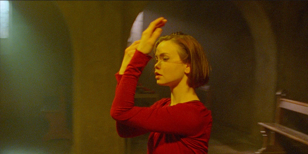 Eine junge Frau (Tatiana Feldman) tanzt in einer Kapelle. Beide Hände hält sie vor ihr Gesicht. Buntes Licht trifft ihr Gesicht. Im Hintergrund fällt Tageslicht durch die Kapellenfenster.