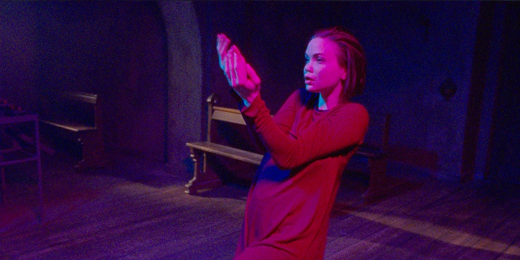 Die Gläubige (Tatiana Feldman) tanzt leicht zurückgelehnt. Magentafarbenes Licht trifft ihr Gesicht und sie trägt ein rotes Kleid. Ihre Haare fallen gleichmäßig nach hinten.