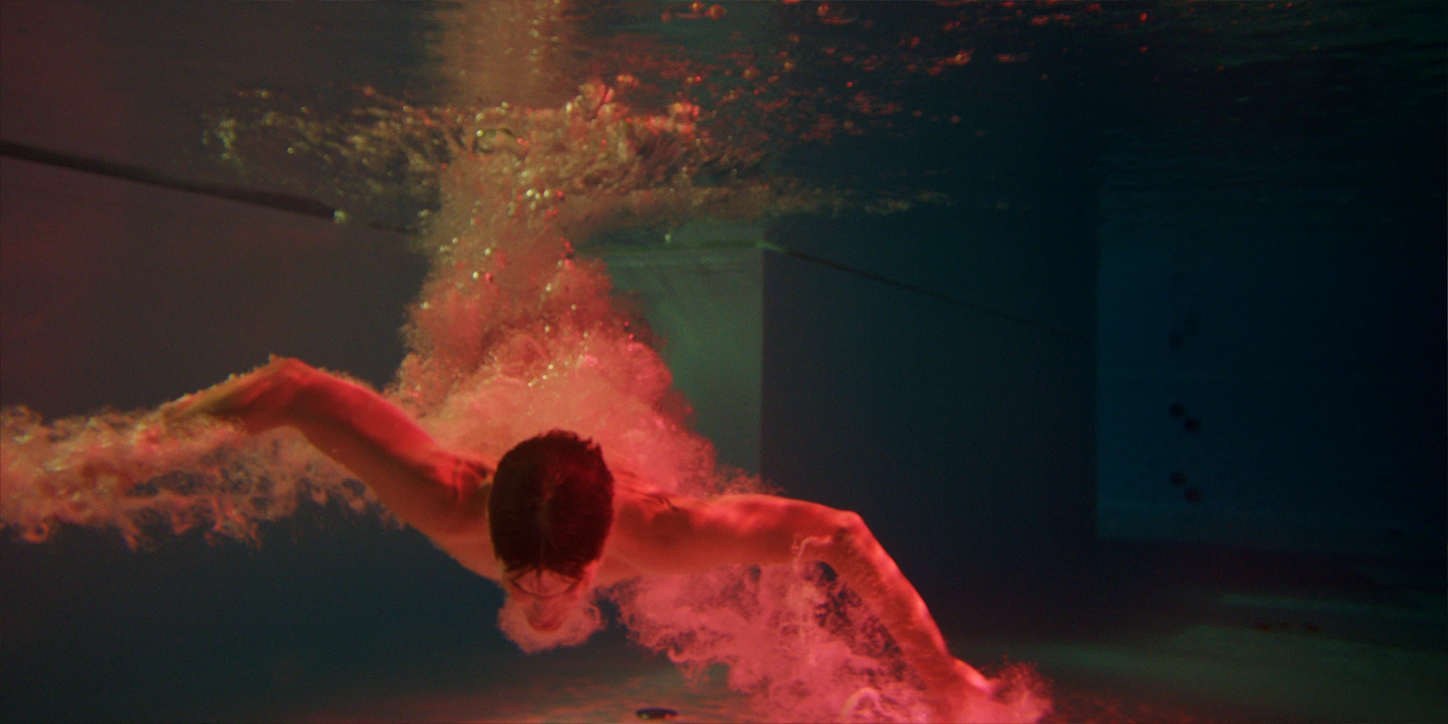 Louis (Benjamin Lillie) taucht mit einem Kopfsprung wie ein Vogel in einen grünlichen Pool ein. Die erzeugten Luftblasen erleuchten rötlich. 