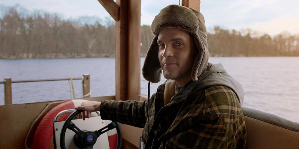 Ein junger Mann (Benjamin Lillie) steuert ein aus Holz gebautes Floß über einen herbstlich anmutenden See. Er trägt eine warme Mütze. Sein Blick ist erwartungsvoll im Werbespot »True Romance«.