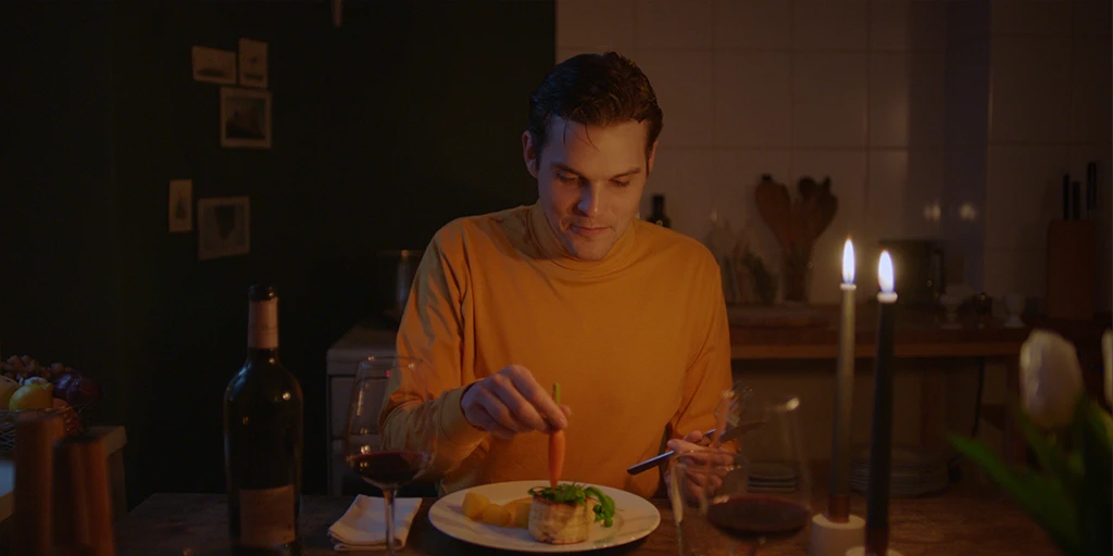 Er (Benjamin Lillie) hat ein stilvolles Abendessen für sie vorbereitet. Er sitzt in gelben Oberteil in seiner Küche. Es steht Wein auf dem Tisch. Er tunkt eine Möhr in eine Mini Quiche im Werbespot »True Romance«.