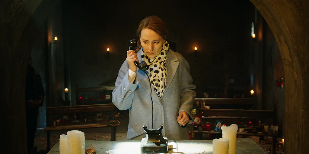 Eine Frau mit blonden Haaren steht vor einem Altar und führt den Hörer eines schwarzen Wählscheibentelefons zu ihrem Ohr. Im Hintergrund sind viele Gedenkkerzen auf den Kirchenbänken aufgestellt. Hinter dem Kapellenbogen ist die Gestalt eines Mannes zu erkennen.