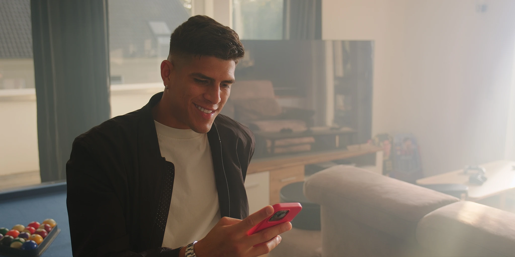 Fußballprofi Piero (Piero Hincapié) sitzt in seiner Villa auf einem Billiardtisch und ließt eine Textnachricht auf seinem Handy. Er trägt eine schlichte schwarze Jacke und ein weißes Shirt. Tiefstehende Sonne flutet den Raum. Im Hintergrund ist ein ausgeschalteter Fernseher zu sehen.