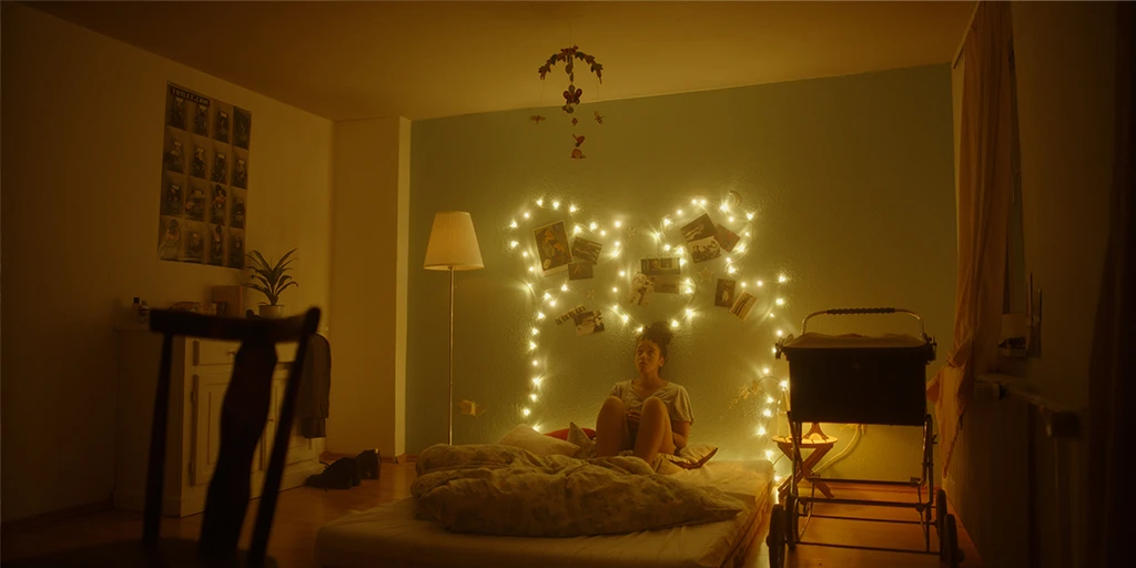Eine junge Frau, Lena (Shari Asha Crosson) sitzt in einem Kinderzimmer. Sie betrachtet ein Mobile. Hinter ihr leuchtet eine Lichterkette. Lena sieht nachdenklich aus. Der Raum ist in warmes Licht getunkt.