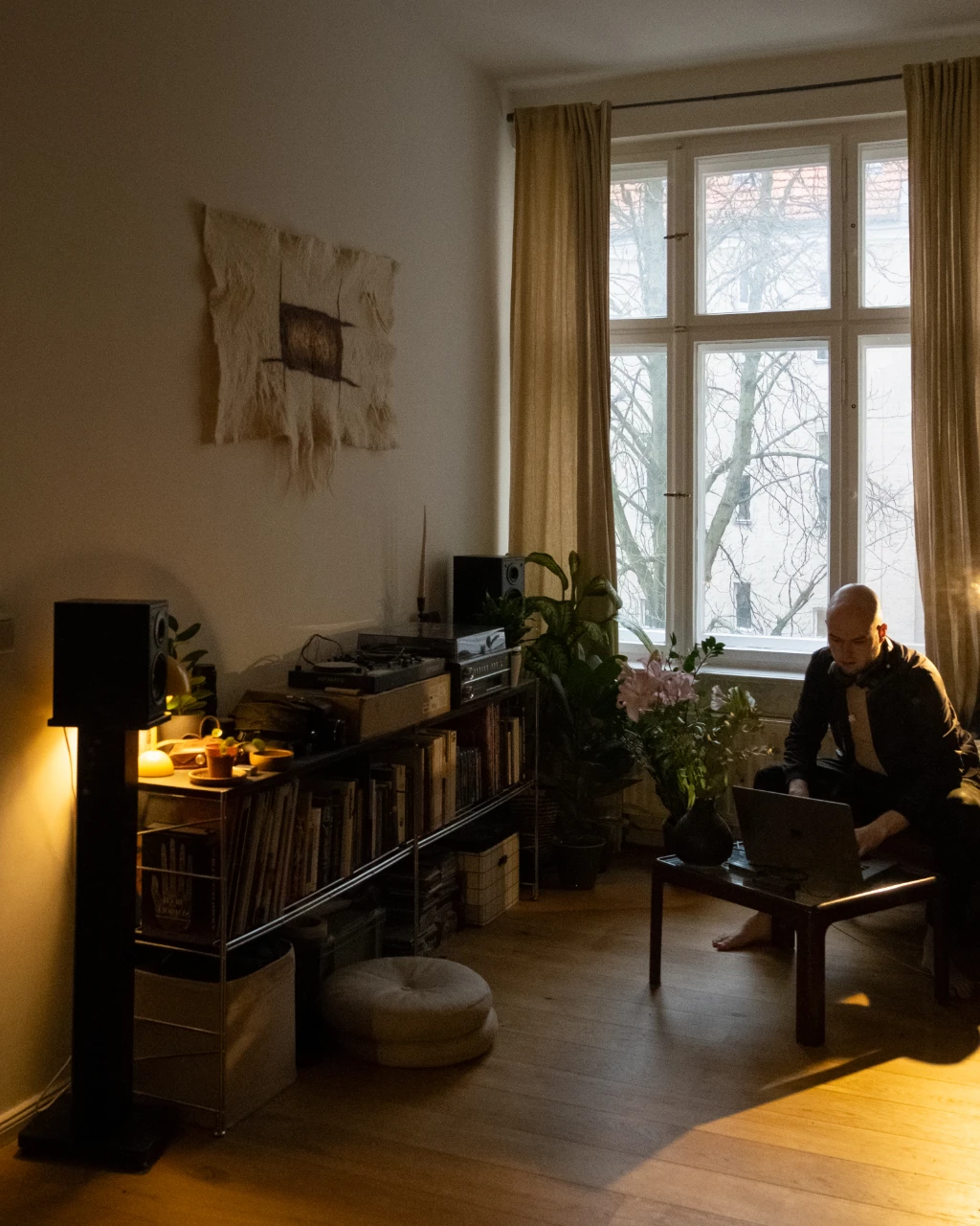 Ein junger Mann sitzt in einem großem Wohnzimmer an seinem Macbook. Im Regal links stehen Schallplatten. Durch das Fenster neben ihm kommt Tageslicht.