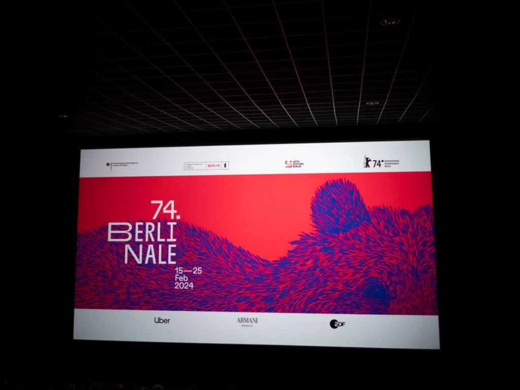 Auf der Leinwand eines Kinos leuchtet ein Introscreen, auf dem ein blauer Bär auf rotem Hintergrund abgebildet ist. Daneben steht geschrieben '74. Berlinale'.