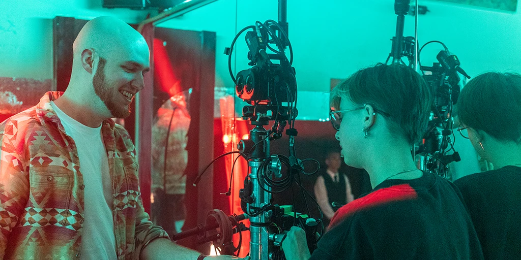 Ein junger Mann und eine junge Frau stehen seitlich vor einem Spiegel. In ihrer Mitte ist eine Virtual Reality Kamera platziert. Beide arbeiten an dem Kamerarig. Der junge Mann lächelt. Die Szene ist in rotes und grünes Licht getaucht.