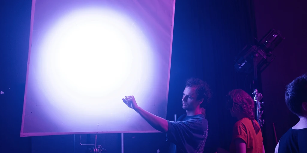 Ein junger Mann steht vor einem 1x1 Meter großem Rahmen, durch den Licht scheint. Er macht die Hand zu einer Faust und prüft das Licht. Neben ihm steht eine junge Frau mit rotem Shirt. Bläuliches Licht durchflutet den Raum.