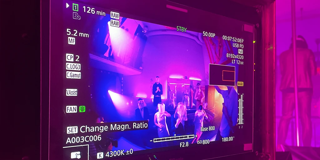 Ein Monitor zeigt einen bläulich-magentafarbenen ausgeleuchteten Stripclub. Tänzer:innen tanzen auf den Tischen.