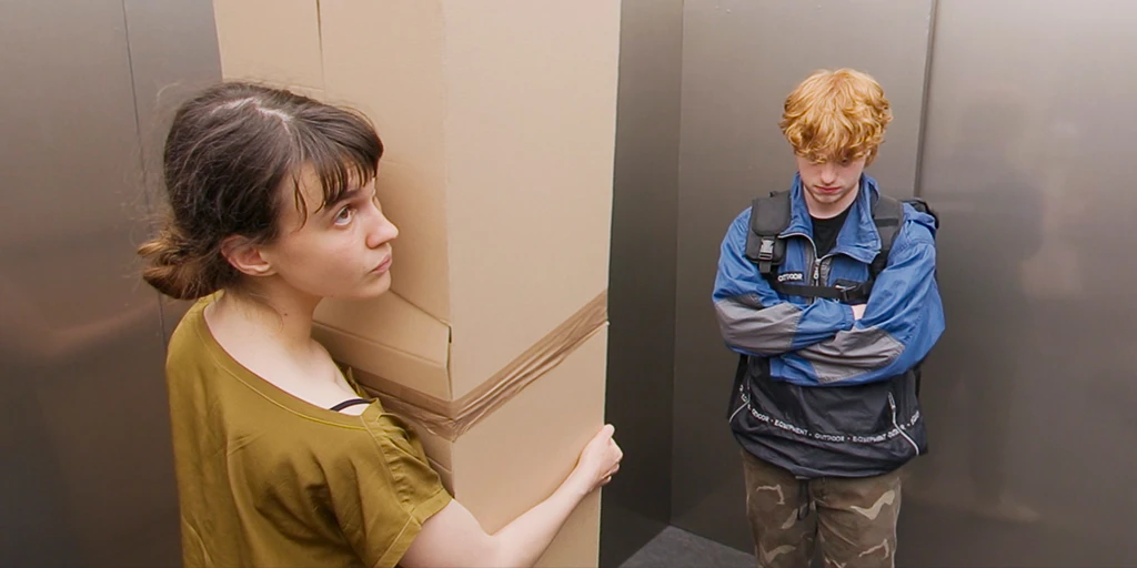 Eine junge Frau steht mit einem groÃŸen Karton in einem Aufzug und schaut zur Decke. Sie hat dunkle Haare, braue Augen und trÃ¤gt ein gelbes Shirt. Rechts neben ihr steht ein junger Mann in blauer Jacke. Er hat rote Haare, trÃ¤gt einen Rucksack und schaut zum Boden.