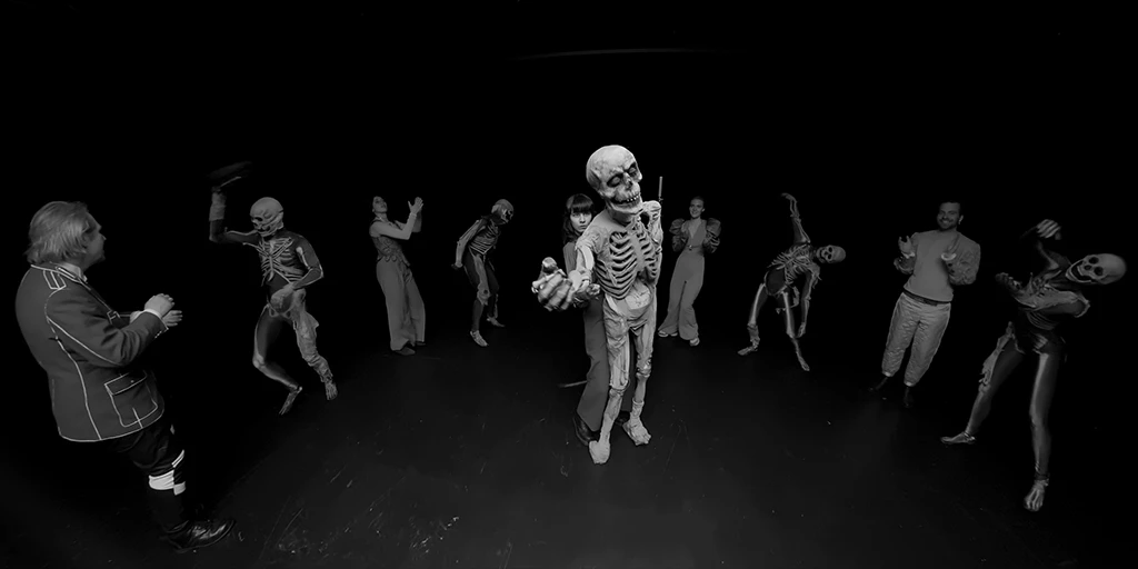 Wir stehen vor einer Puppenspielerin mit dunklen Haaren, die eine Skelett-Puppe steuert und eine einladende Geste macht. Um sie herum tanzen insgesamt 8 Menschen einen Totentanz. Dabei tragen sie unter anderem SkelettkostÃ¼me.