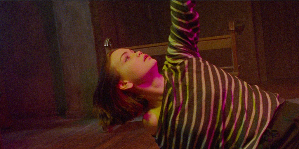 Eine junge Frau (Tatiana Feldman) bewegt sich in einer Tanzpose in einer Kapelle. Ihr linker arm ist nach oben gestreckt. gelb-magentafarbendes Licht trifft ihr Gesicht.