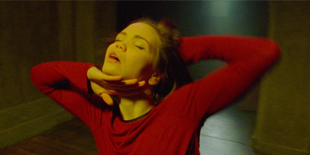 Eine junge Frau (Tatiana Feldman) tanzt in einem roten Kleid in einer Kapelle. Dabei streicht ihre rechte Hand unter ihrem Kinn entlang, ihre linke Hand ist hinter ihrem Kopf.